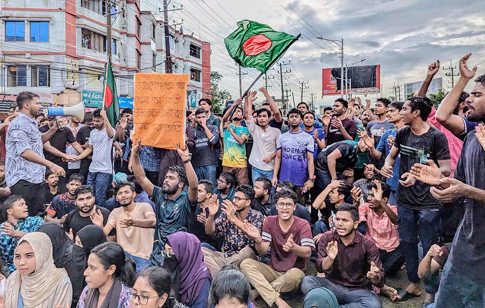 Youth protesters gather in street demonstration, some seated, some standing, waving national flag