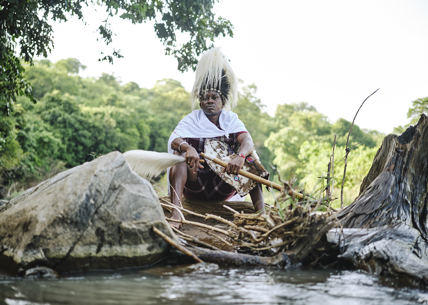 Tharaka spiritual leader in ceremonial headdress sits in dugout canoe by riverbank, Kenya