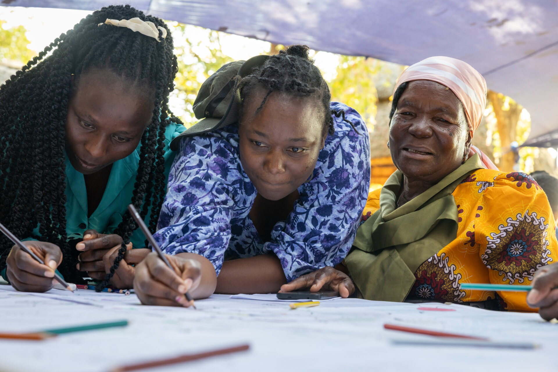Three generations gather under shade, drawing and mapping together with colored pencils