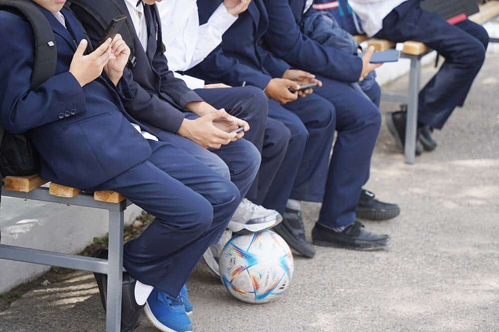 Students in navy uniforms sit on a bench with smartphones, a soccer ball at their feet